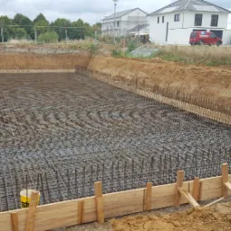 Szeroki wykop pod fundamenty, zabezpieczony drewnianym szalunkiem i wypełniony siatką zbrojeniową, w tle budowa domów jednorodzinnych.