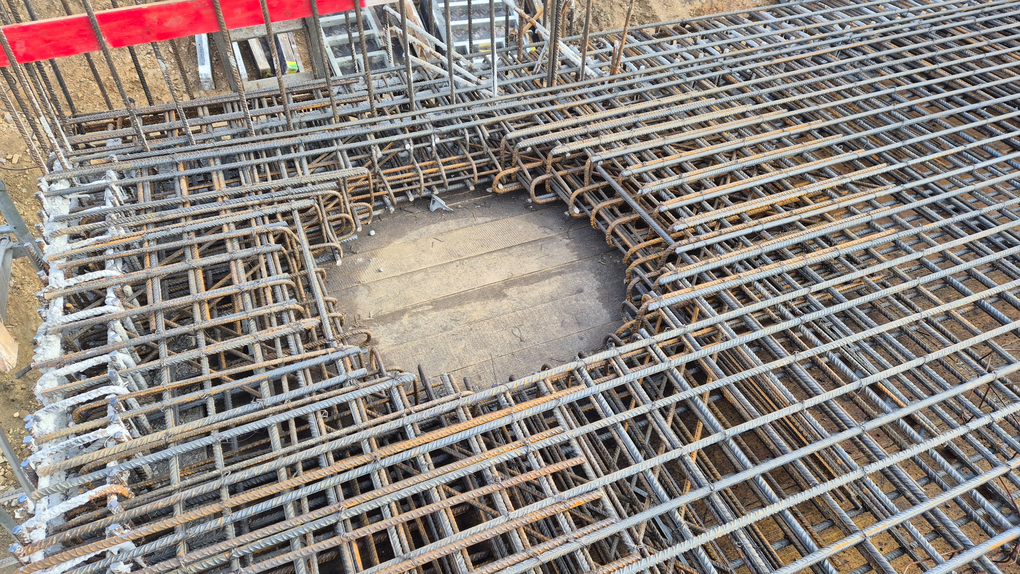Siatka z prętów zbrojeniowych ułożona na deskowaniu, przygotowana do zalania betonem, widok z góry, fragment czerwonej barierki ochronnej w tle.