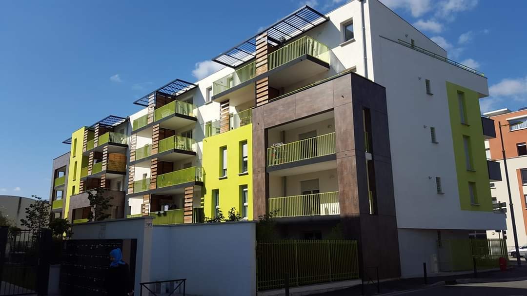 Nowoczesny budynek wielorodzinny z balkonami o limonkowych barierkach, elewacja w kolorach bieli, zieleni i brązu, widok od dołu w słoneczny dzień.