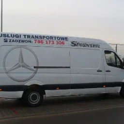 Biały bus Mercedes Sprinter z napisem 'Usługi Transportowe' i numerem telefonu, zaparkowany na zewnątrz. Widok z boku, lekko z przodu. Szary asfalt i budynki w tle.
