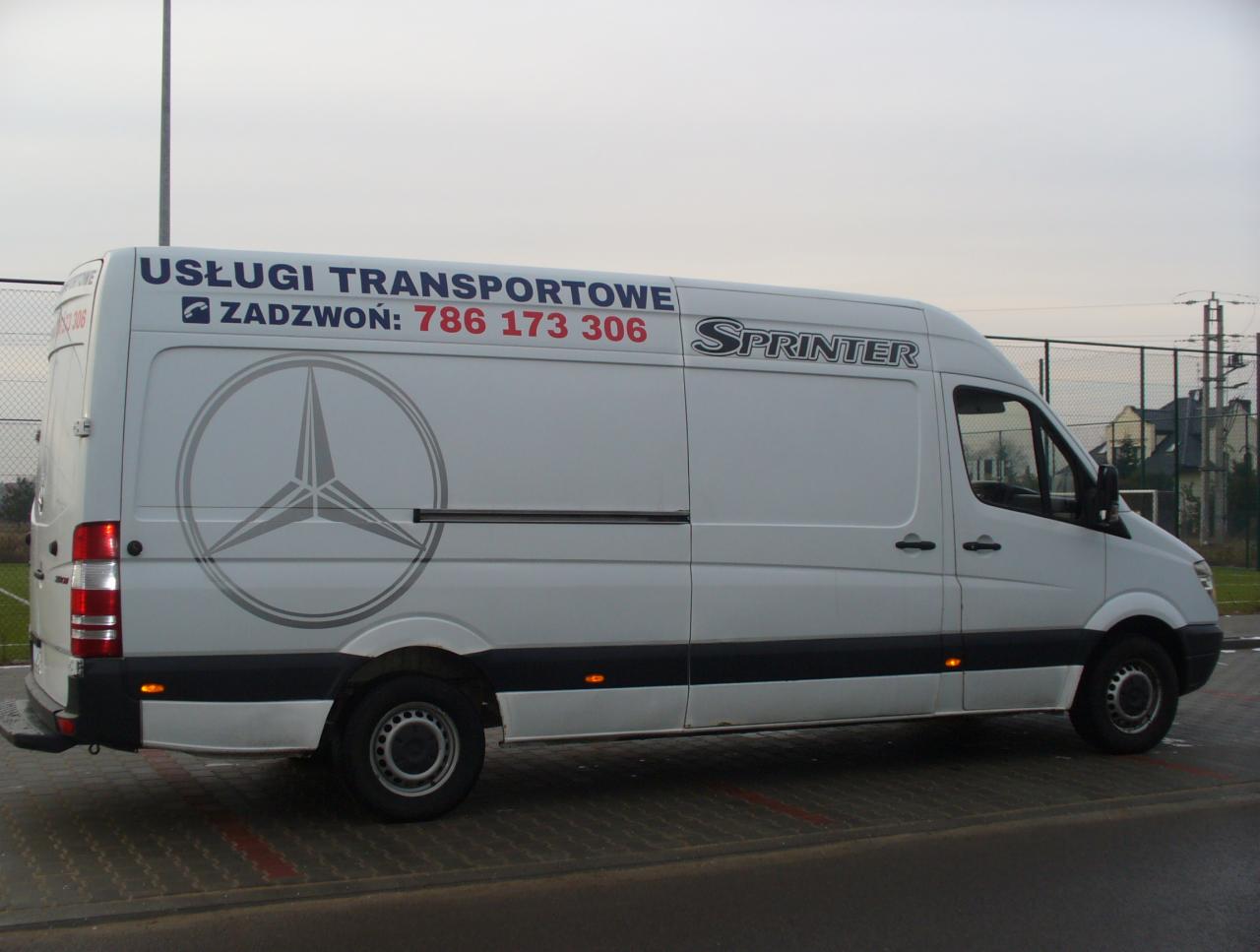 Biały bus Mercedes Sprinter z napisem 'Usługi Transportowe' i numerem telefonu, zaparkowany na zewnątrz. Widok z boku, lekko z przodu. Szary asfalt i budynki w tle.
