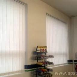 Jasne żaluzje pionowe w biurze, widoczne z boku, z ekspozytorem taśm izolacyjnych w tle. Oświetlenie dzienne, neutralna kolorystyka ścian.