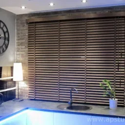 Kuchnia z drewnianymi żaluzjami poziomymi, widok na okno z blatem i zlewozmywakiem, dekoracyjny zegar na ścianie, oświetlenie LED pod szafkami.