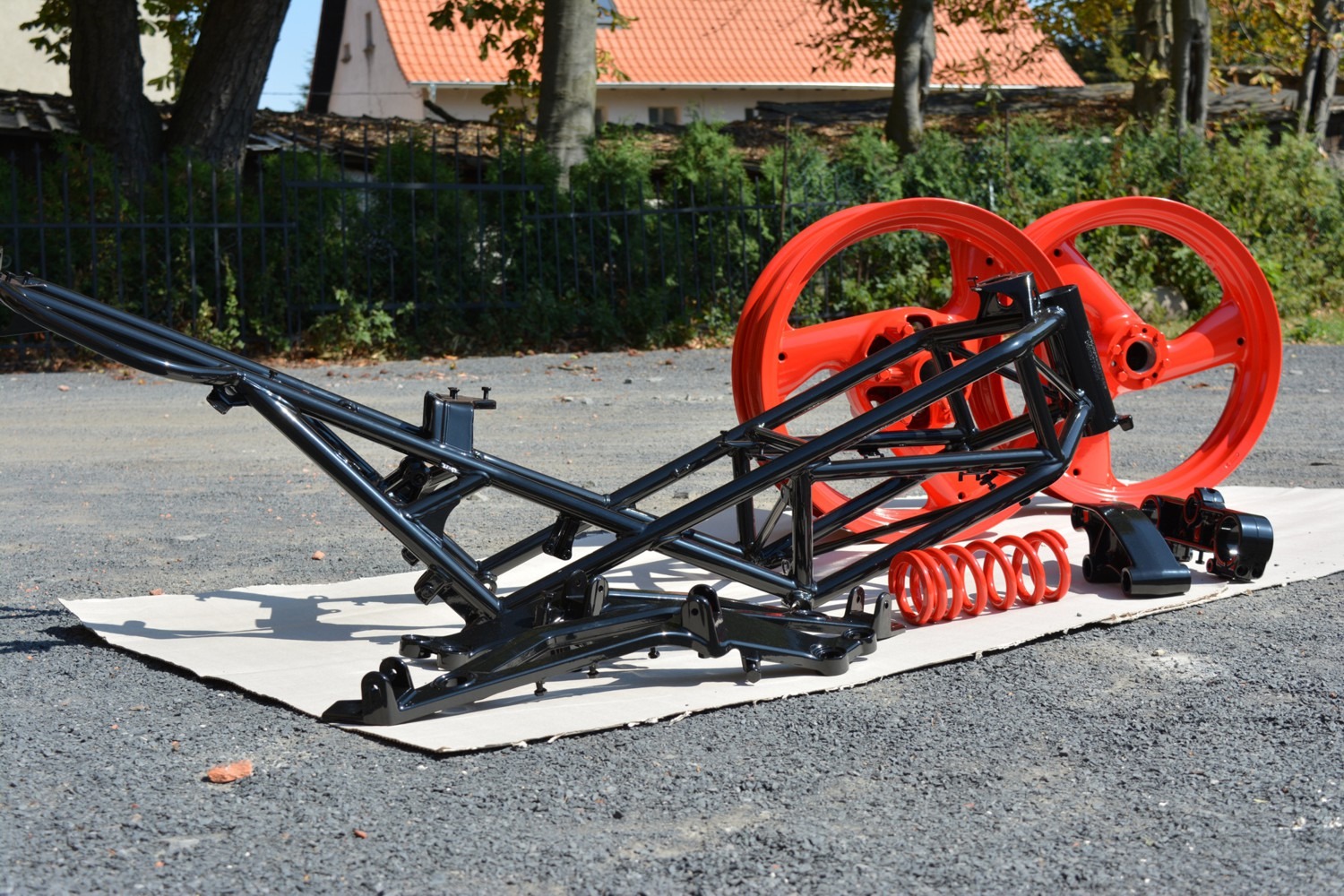 Części motocyklowe po renowacji: czarna rama, czerwone felgi i sprężyna, ułożone na kartonie na zewnątrz.