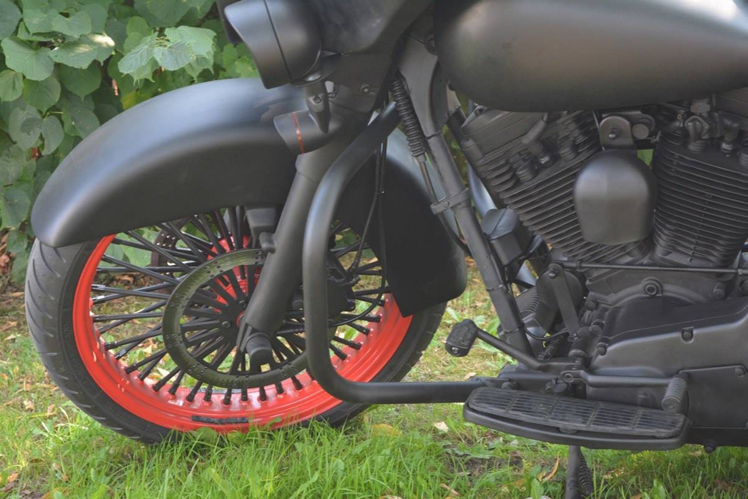 Matowy czarny motocykl z czerwonymi akcentami na obręczy koła, widoczne detale silnika i przedniego zawieszenia na tle zielonej trawy i krzewów.