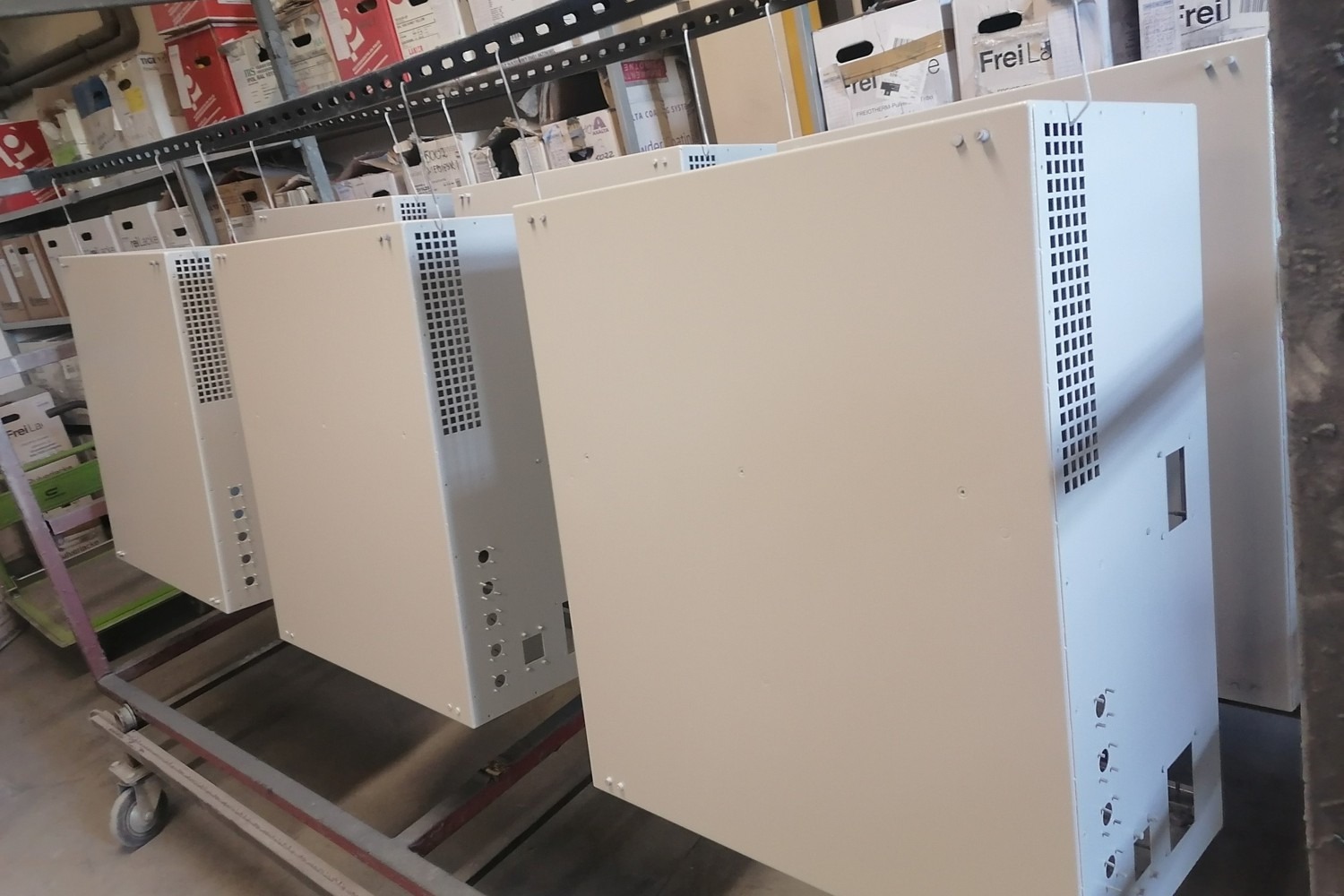 Białe metalowe obudowy z perforacją, zawieszone na wieszakach, prawdopodobnie po procesie malowania, widoczne ślady po zawieszeniu, tło magazynowe z kartonami.