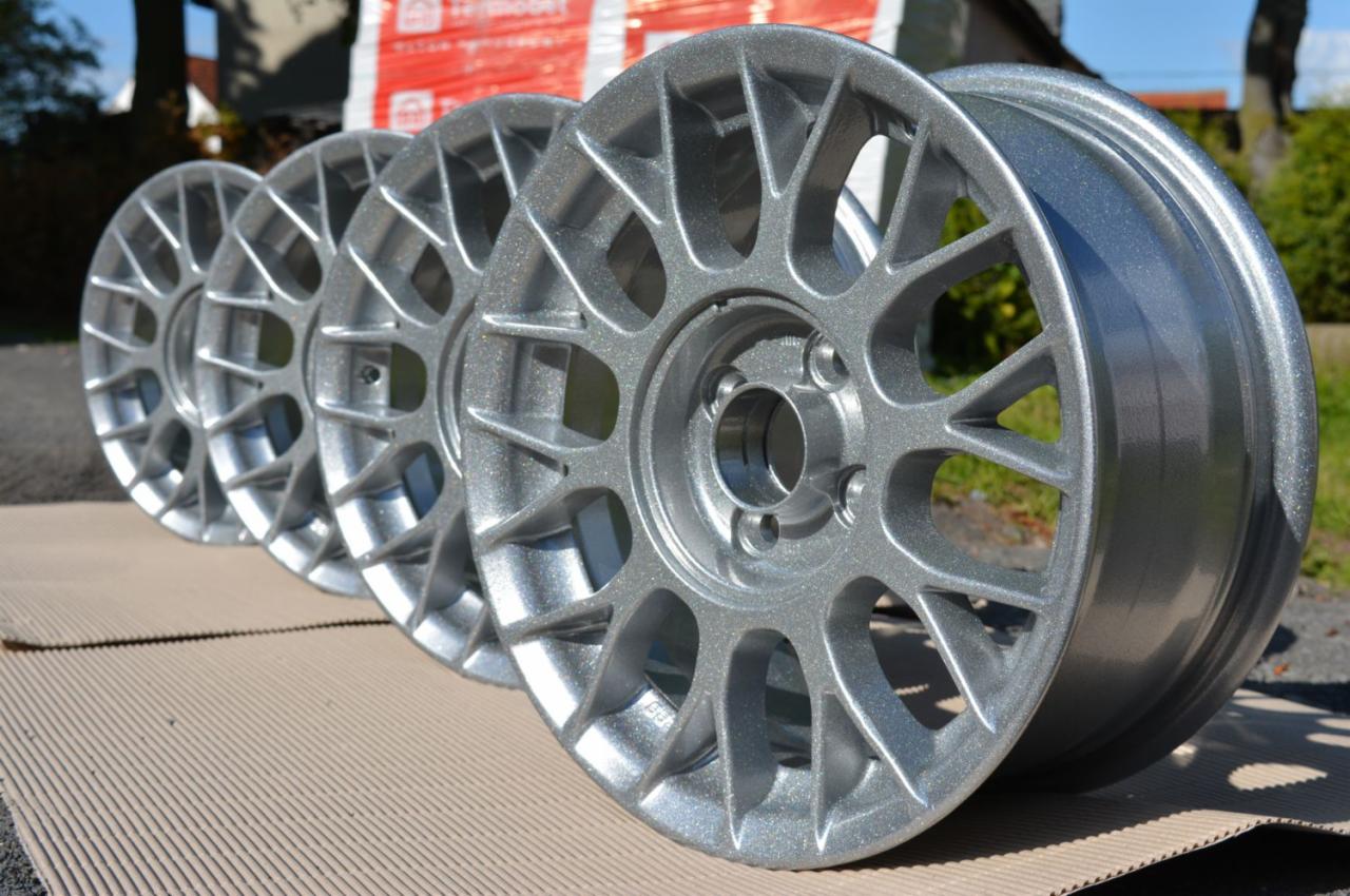 Cztery felgi aluminiowe po renowacji, pokryte srebrnym, metalicznym lakierem, ustawione na tekturowej powierzchni na zewnątrz.