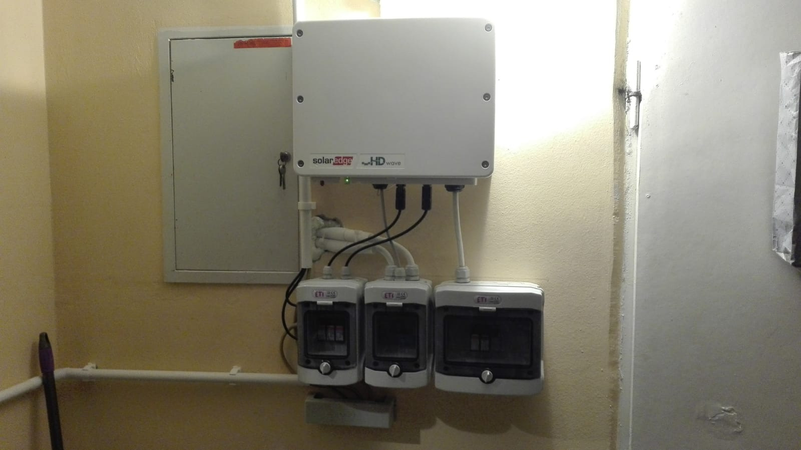 Biały inwerter solarny z trzema zabezpieczeniami ETI zamontowany na żółtej ścianie, widoczna szafka elektryczna z kluczem i rura grzewcza w tle.