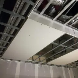 Montaż sufitu podwieszanego z płyt gipsowo-kartonowych na metalowej konstrukcji nośnej, widoczne instalacje wentylacyjne powyżej.