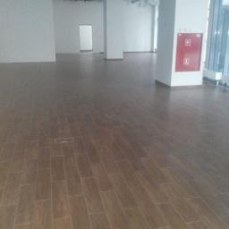 Usługi remontowo-wykończeniowe - Ułożona, brązowa podłoga z płytek imitujących drewno w przestronnym, pustym pomieszczeniu biurowym. Widoczne kolumny i instalacje pod sufitem.