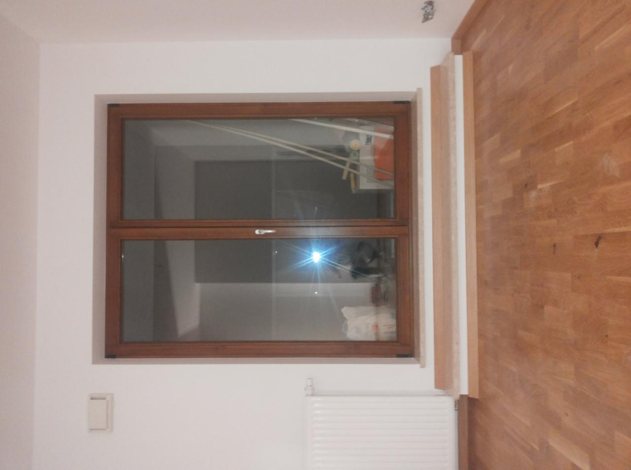 Drewniane okno z widocznym grzejnikiem i fragmentem drewnianej ściany w jasnym wnętrzu. Częściowo widoczne listwy przypodłogowe i włącznik światła.