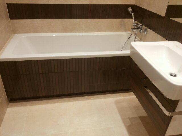 Nowoczesna łazienka z wanną obudowaną ciemnym drewnem i umywalką. Beżowe płytki podłogowe i pasy dekoracyjne na ścianach. Bateria wannowa z słuchawką.