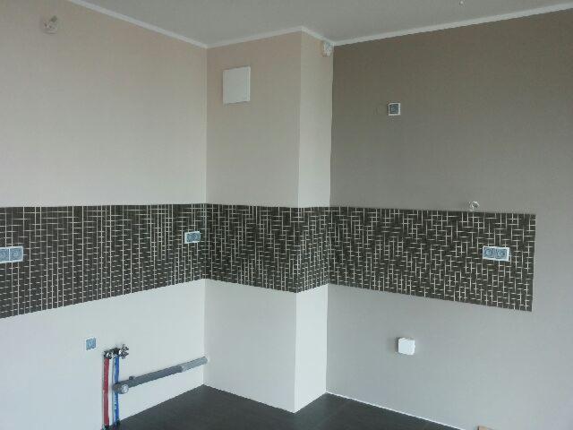 Wykończona ściana z czarnymi, wzorzystymi płytkami ułożonymi w pasie, z widocznymi gniazdkami i instalacją wodną w wyremontowanej kuchni. Ściany w odcieniach beżu i szarości.