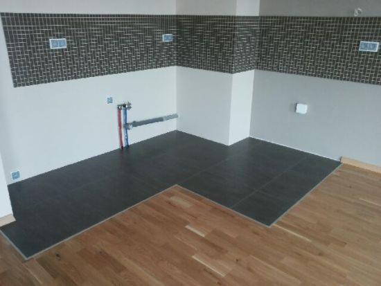 Połączenie dwóch typów podłóg: ciemne płytki i jasny parkiet, na ścianach mozaika. Widoczne rury instalacji wodnej. Etap prac wykończeniowych.