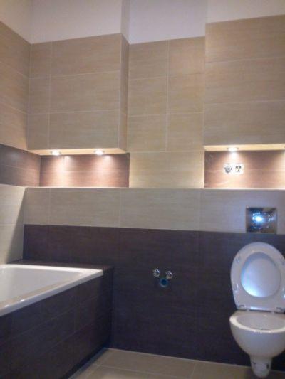 Wykończona łazienka z beżowo-brązowymi płytkami, oświetleniem wnękowym i otwartą klapą sedesu. Minimalistyczny design z widoczną wanną.