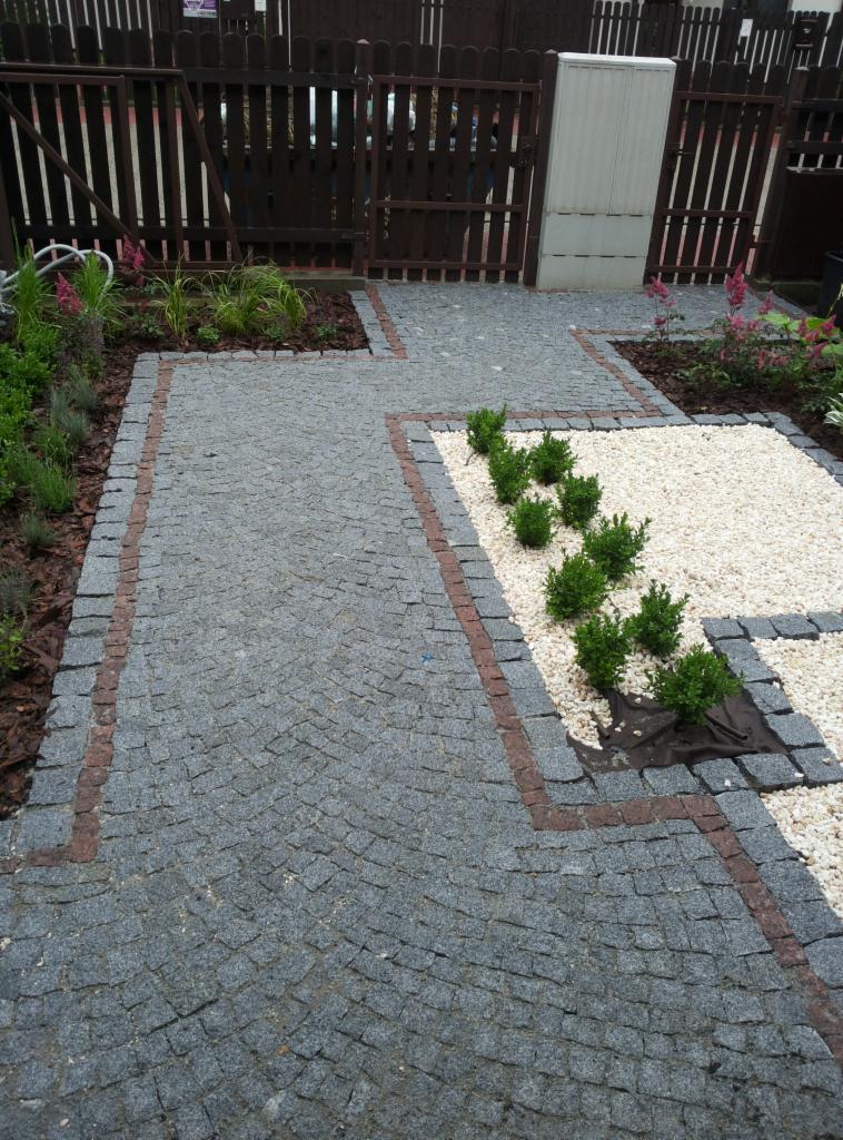 Granitowa kostka brukowa ułożona w łukowaty wzór, obramowana czerwoną kostką. Biały żwir i młode krzewy. Drewniany płot w tle.