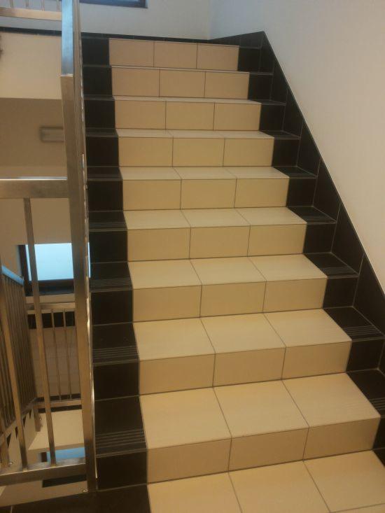 Schody wyłożone beżowymi płytkami z czarnymi krawędziami, widok z dołu, metalowa balustrada po lewej stronie. Jasne, neutralne tło ścian.