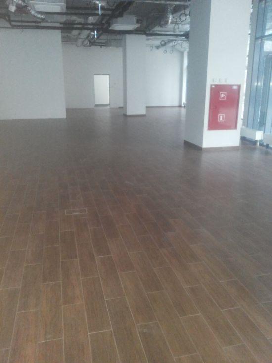 Ułożona, brązowa podłoga z płytek imitujących drewno w przestronnym, pustym pomieszczeniu biurowym. Widoczne kolumny i instalacje pod sufitem.