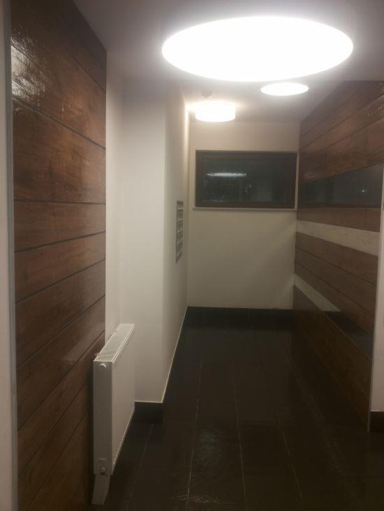 Korytarz z drewnianą boazerią, błyszczącą podłogą z płytek i okrągłymi lampami sufitowymi. Detal: biały grzejnik przy ścianie. Nowoczesne wykończenie wnętrza.