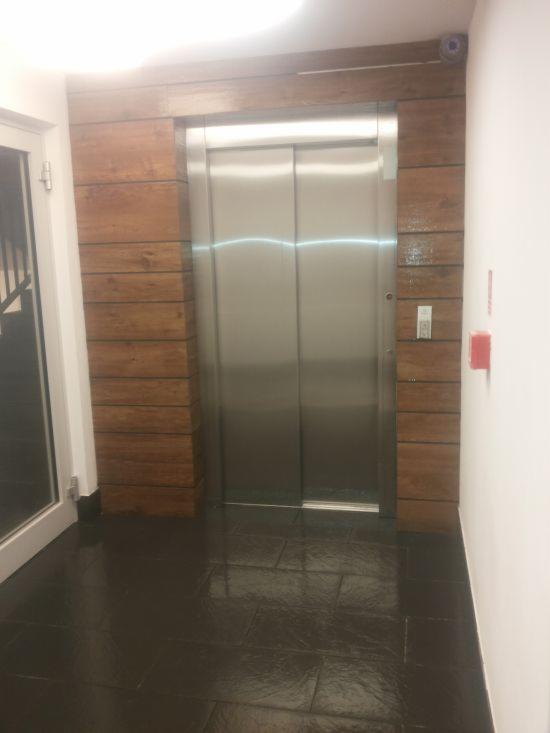 Wykończenie ściany z drewnianych paneli wokół windy, uzupełnione o czarną, błyszczącą posadzkę. Widoczny fragment drzwi i biała ściana z elementami przeciwpożarowymi.