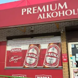 Fasada sklepu z alkoholem z czerwonym szyldem 'PREMIUM ALKOHOLE' i logo 'WARKA'. Na ścianie reklama piwa Warka Classic z informacją o zawartości alkoholu w piwie.