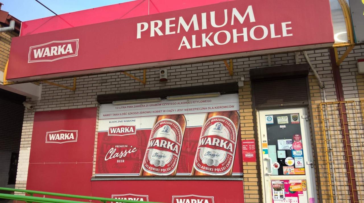 Fasada sklepu z alkoholem z czerwonym szyldem 'PREMIUM ALKOHOLE' i logo 'WARKA'. Na ścianie reklama piwa Warka Classic z informacją o zawartości alkoholu w piwie.