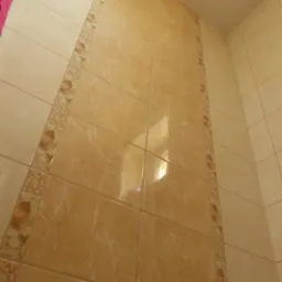 Beżowe płytki ścienne w łazience, ułożone pionowo z dekoracyjnym pasem po bokach, widoczne pod kątem. Na wieszaku fragment różowego ręcznika.