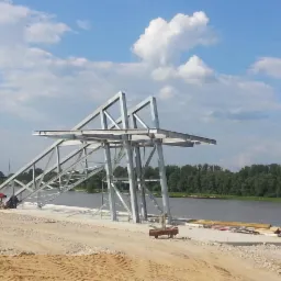 Konstrukcja stalowa w trakcie montażu nad rzeką, z pracownikami w oddali i błękitnym niebem w tle. Widoczne elementy rusztowań i narzędzia.