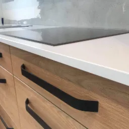 Fragment kuchni z białym blatem i drewnianymi szafkami z czarnymi, długimi uchwytami, płyta indukcyjna wkomponowana w blat, ściana w trakcie wykończenia.