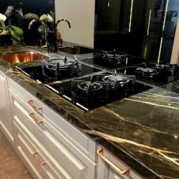 Czarny blat z marmuru ze złotymi żyłami, wbudowana czarna płyta gazowa z czterema palnikami, szare szafki kuchenne z miedzianymi uchwytami, miedziany zlew, kwiaty w wazonie.