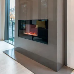 Nowoczesny kominek elektryczny w obudowie z szarego betonu architektonicznego, z odbiciem wnętrza salonu w czarnej, szklanej ramie.