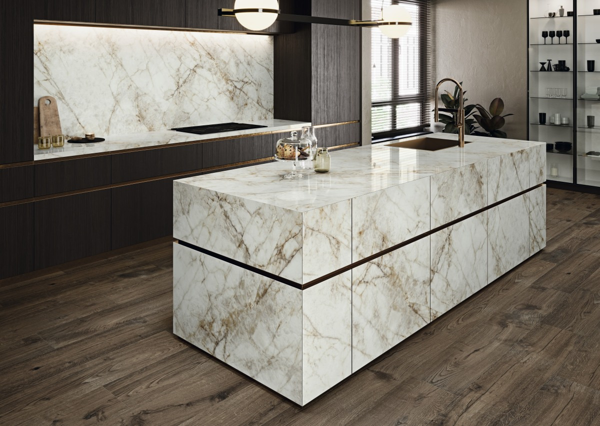 Wnętrze kuchni z wyspą pokrytą białym marmurem z brązowymi żyłami, pasującym do blatu za nią; ciemne drewniane szafki z brązowymi akcentami, drewniana podłoga, nowoczesne oświetlenie.