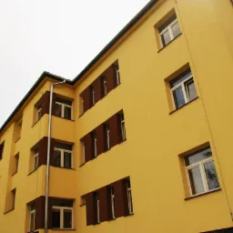 Żółty budynek wielorodzinny po dociepleniu elewacji, z brązowymi obramowaniami okien i rynnami, sfotografowany z dołu pod kątem.