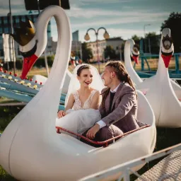 Uśmiechnięta para młoda w strojach ślubnych siedzi w białym łabędziu na karuzeli, patrząc na siebie z czułością, z budynkami w tle.