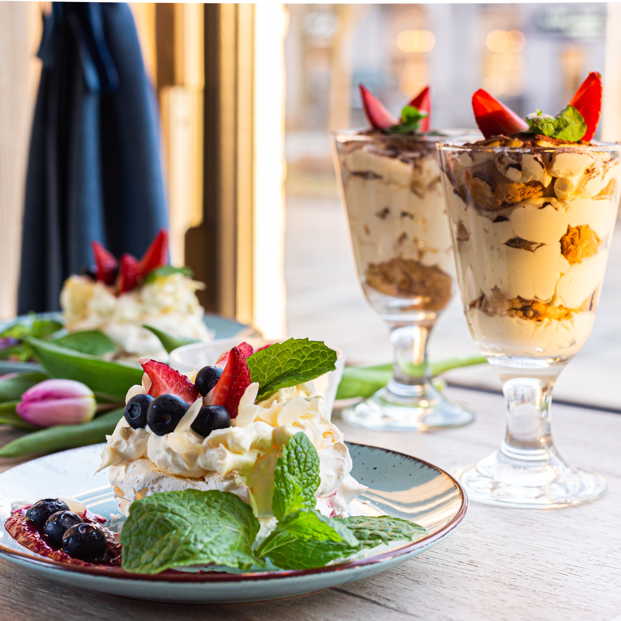 Pavlova z bitą śmietaną, truskawkami, borówkami i miętą na turkusowym talerzu, obok tulipan i tiramisu w szklance.