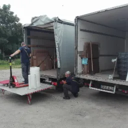 Dwaj pracownicy firmy transportowej przy dwóch otwartych samochodach dostawczych; jeden z nich obsługuje wózek paletowy z folią stretch, drugi kontroluje oświetlenie pojazdu.