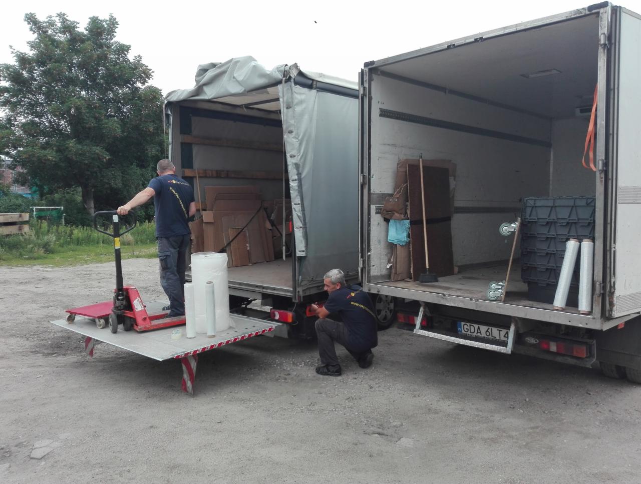 Dwaj pracownicy firmy transportowej przy dwóch otwartych samochodach dostawczych; jeden z nich obsługuje wózek paletowy z folią stretch, drugi kontroluje oświetlenie pojazdu.