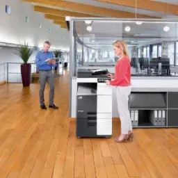 Kobieta obsługuje nową kserokopiarkę w biurze; mężczyzna z tabletem w tle, drewniana podłoga.