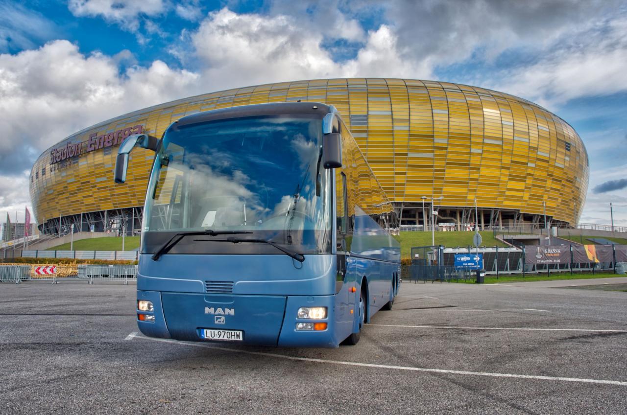 Niebieski autokar MAN na parkingu przed Stadionem Energa Gdańsk, chmury odbijają się w szybie przedniej.