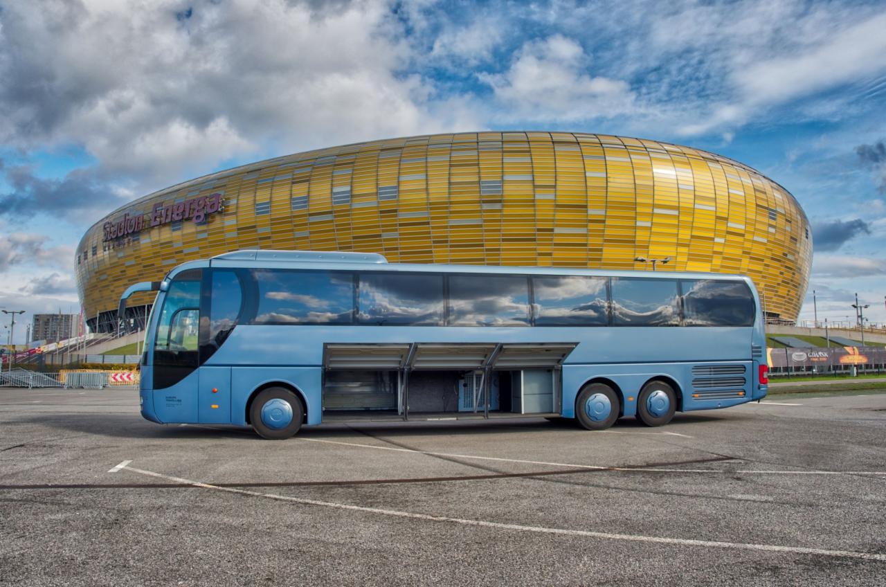 Niebieski autokar turystyczny z otwartymi lukami bagażowymi na parkingu, w tle stadion Energa w Gdańsku.
