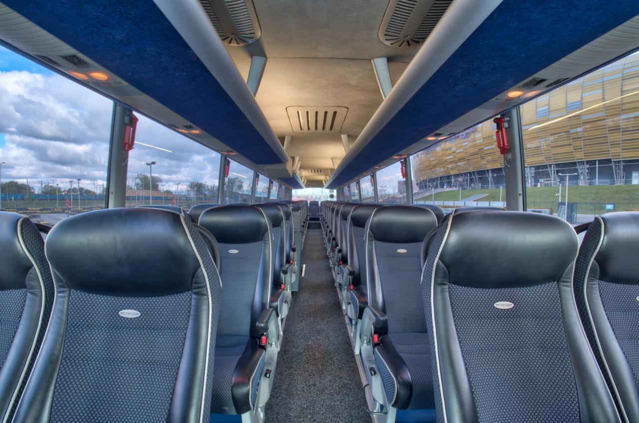 Wnętrze autobusu turystycznego z rzędami czarno-granatowych foteli z zagłówkami, widok na okna i budynek w tle.