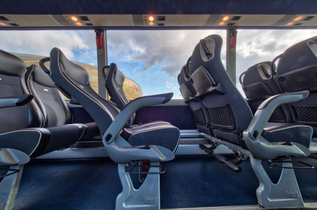 Wnętrze autobusu z widokiem na wygodne fotele pasażerskie z regulowanymi podłokietnikami i siatkami na drobiazgi, za oknem budynek i błękitne niebo.