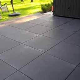 Nowoczesny taras z ciemnoszarej, kwadratowej kostki betonowej, przylegający do ściany budynku. W tle trawnik i meble ogrodowe pod pokrowcem.