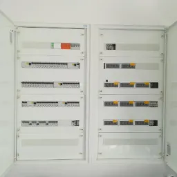 Otwarta, biała szafa rozdzielcza z widocznymi zabezpieczeniami, bezpiecznikami i modułami elektrycznymi, zamontowana na ścianie.