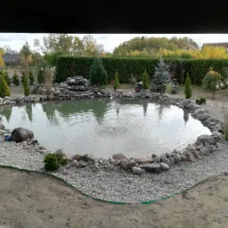 Oczko wodne z fontanną otoczone kamieniami i żwirem, w tle zielony żywopłot i drzewa.