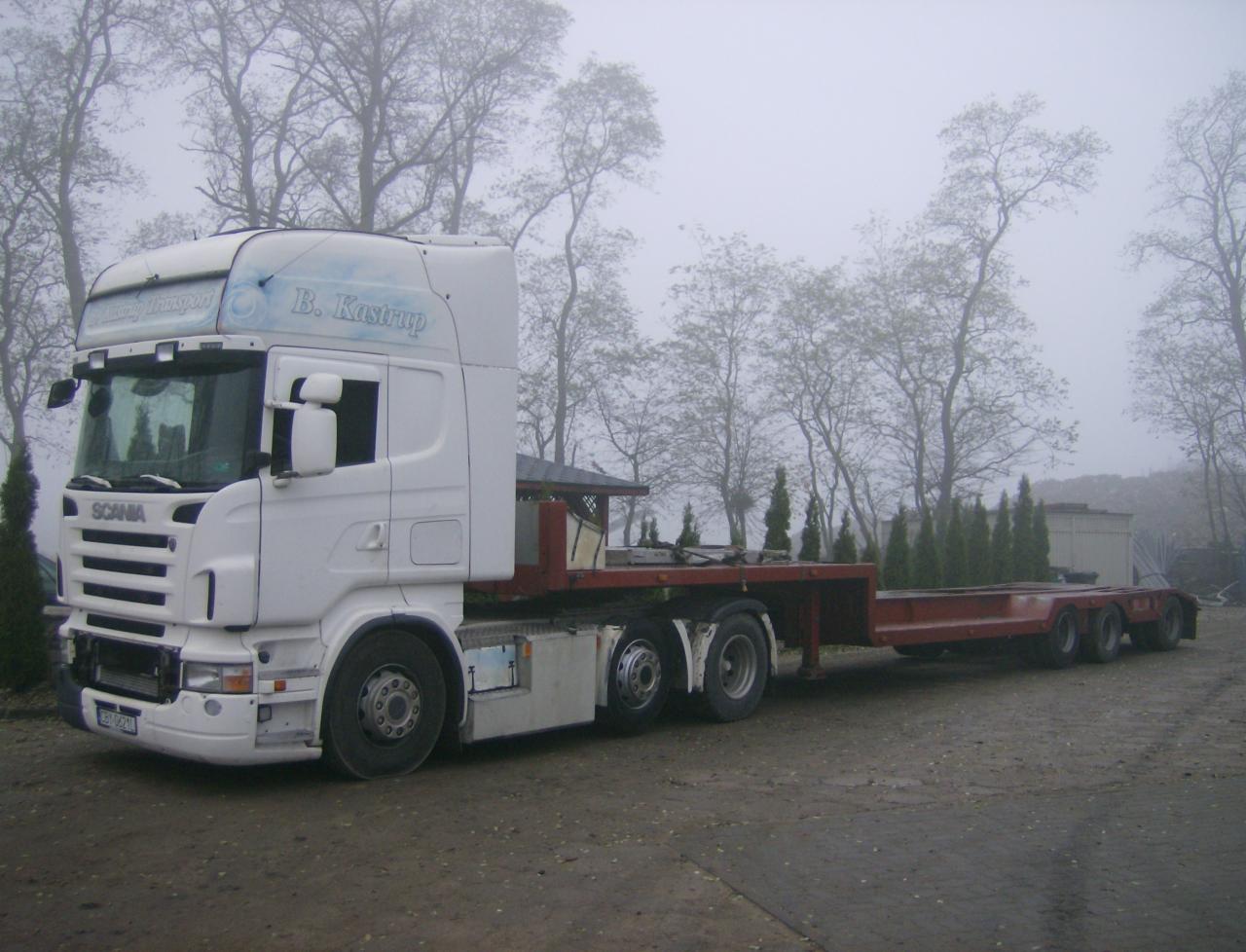 Biała ciężarówka Scania z naczepą niskopodwoziową na placu, w tle drzewa we mgle. Widoczny napis 'B. Kastrup Transport'.