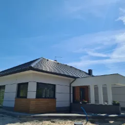 Parterowy dom jednorodzinny z płaskim dachem i białymi ścianami, częściowo wykończony drewnianą okładziną, widoczny w słoneczny dzień na tle błękitnego nieba.