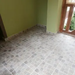 Wykończony balkon z szaro-beżową, wzorzystą terakotą na podłodze, listwy przypodłogowe dopasowane do płytek, drewniana balustrada, zielone ściany i widok na zieleń.