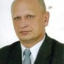 Portret mężczyzny w garniturze i krawacie w odcieniach niebieskiego, z łagodnym uśmiechem i krótkimi włosami, na neutralnym tle.