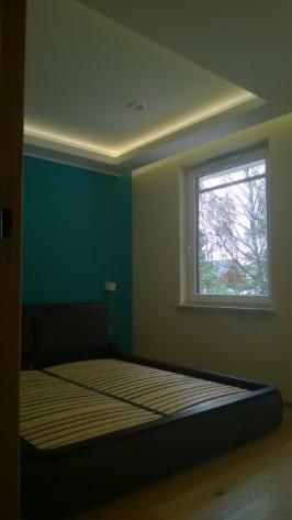 Sypialnia po remoncie: turkusowa ściana, podświetlany sufit i łóżko z drewnianym stelażem. Widok na okno z drzewem na zewnątrz. Minimalistyczny styl.